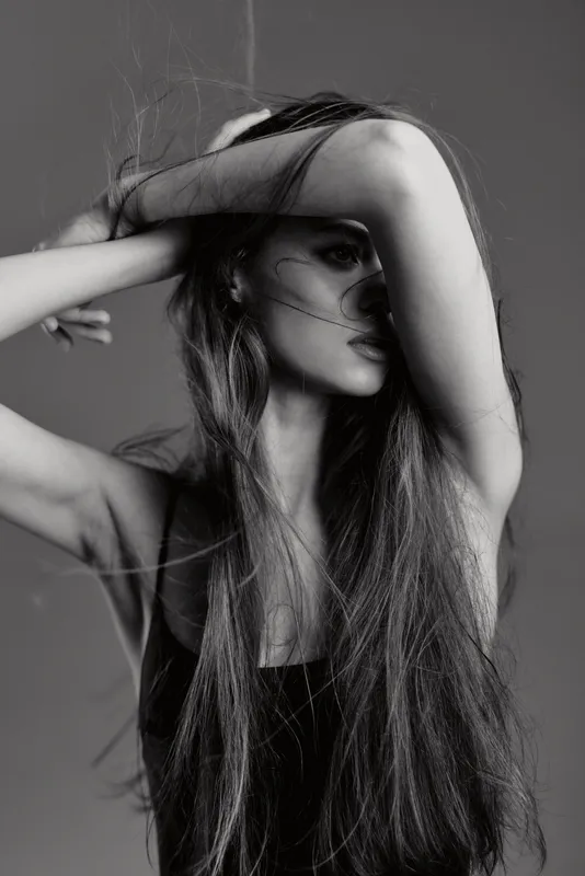Black and white portrait of a woman with long hair posing with arm covering face in studio