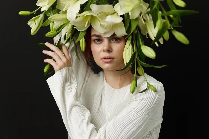 Young woman with floral arrangement on her head in a white outfit, expressing calmness and natural beauty against a dark background, highlighting floral concepts