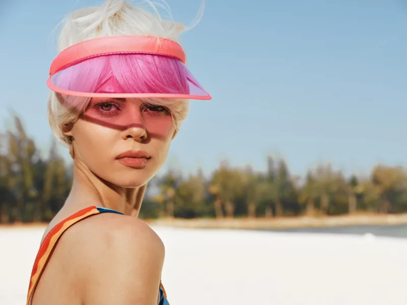 Close up portrait of a stylish woman on a sunny beach, wearing a pink visor and colorful accessories, blonde hair and radiant skin under bright light.