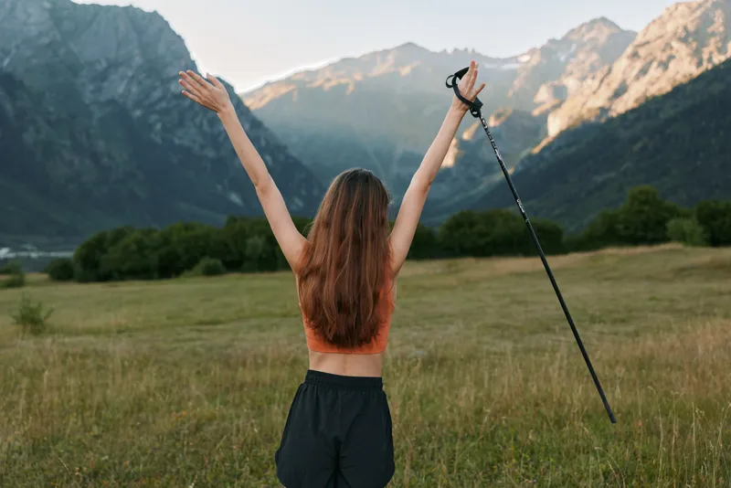 A athletic woman hiker stands in a wide meadow outdoors, arms raised, gripping a trekking pole, radiating freedom and joy as distant mountains glow under a clear sky.
