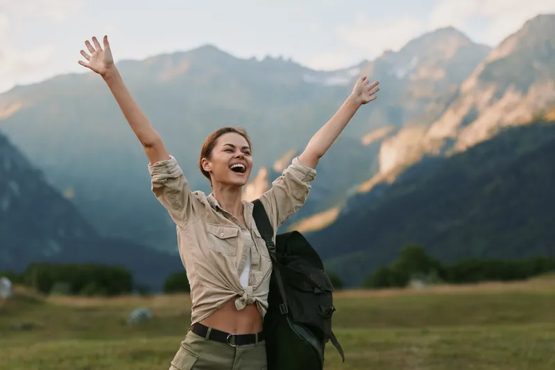 Happy woman outdoors raising hands, smiling with eyes closed, casual outfit, mountain landscape background, sunny day, carefree and joyful mood