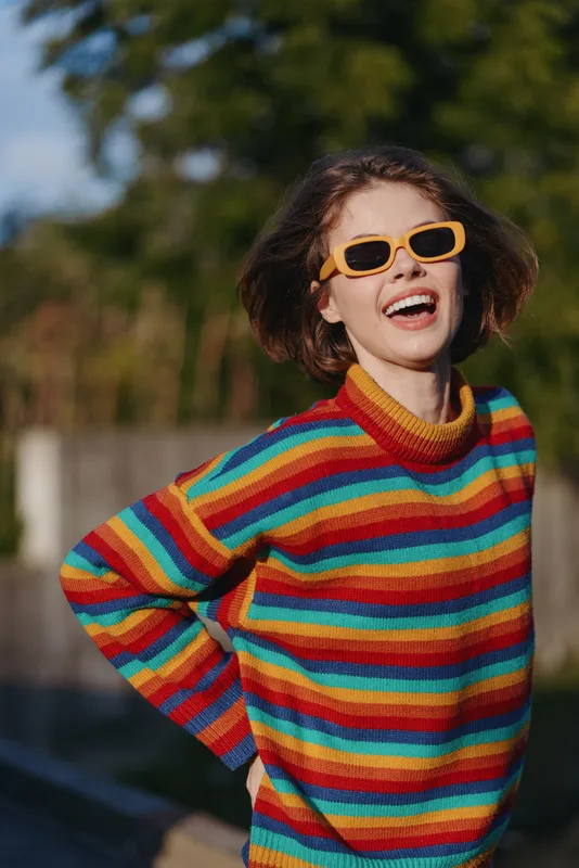 Woman smiling in a striped sweater and orange sunglasses outdoors, young model in her twenties wearing a casual outfit, playful pose with hands on hip, colorful travel lifestyle portrait.
