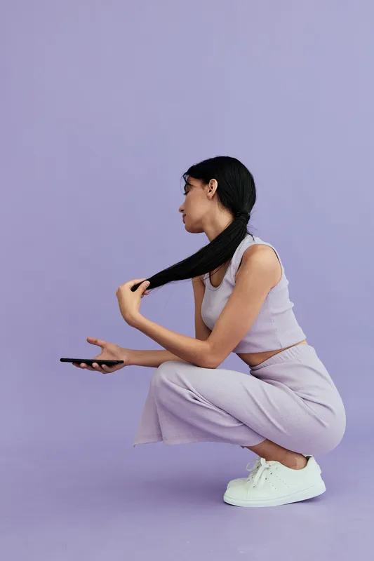 Young woman squatting while holding her long hair and smart phone against a soft purple background, wearing casual light purple outfit and white sneakers.