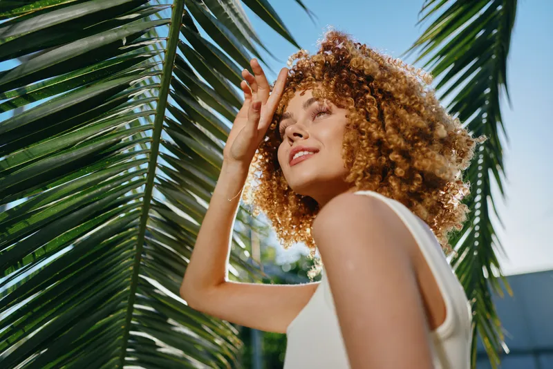 Smiling woman with curly hair enjoys sunlight under palm leaves, wearing white sleeveless top, vibrant outdoor summer scene with clear blue sky background.