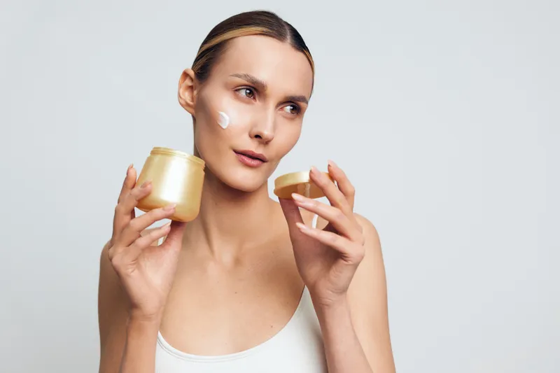 Young woman with clear skin applies face cream from a jar while looking thoughtfully to the side against a plain light background.