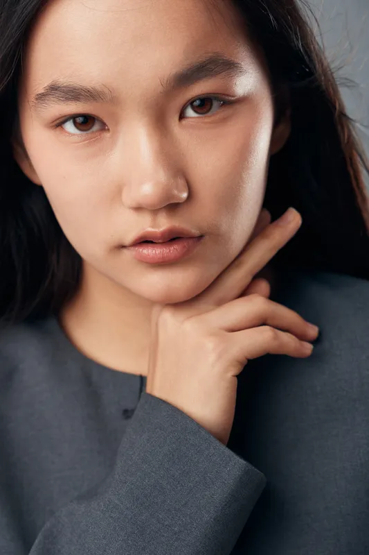 Portrait of young woman with natural makeup and dark hair posing with hand near face wearing gray clothing studio background. Beauty expression and fashion model closeup with soft lighting and