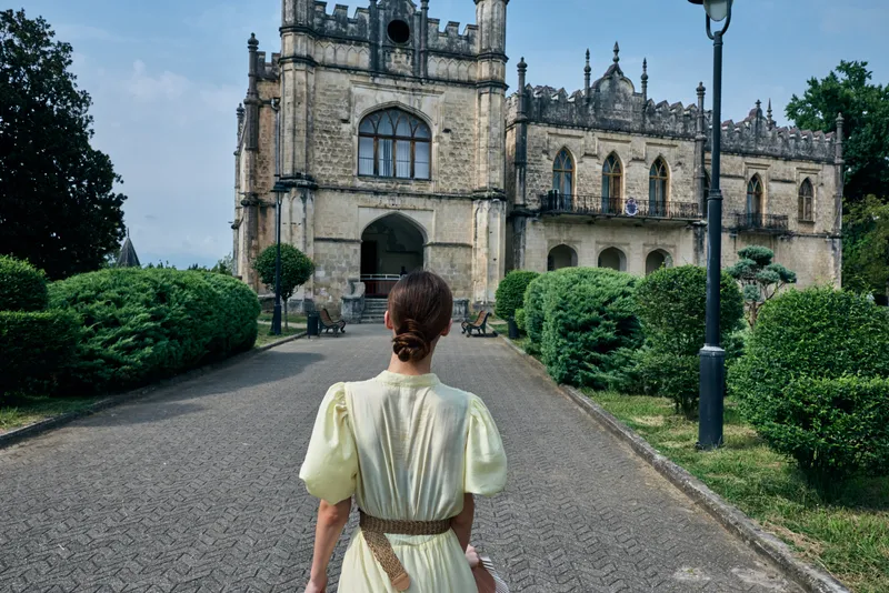 A woman in a pale yellow vintage dress walks toward a grand historic castle along a tree lined path and manicured garden surroundings.