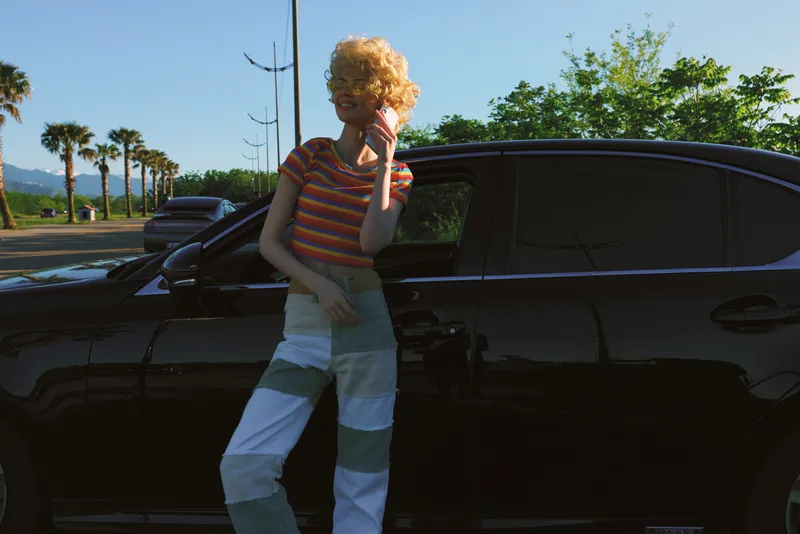 Young woman with curly orange hair leans on a dark SUV on a sunny street, exuding retro film color and casual street style as the moment feels candid and confident.