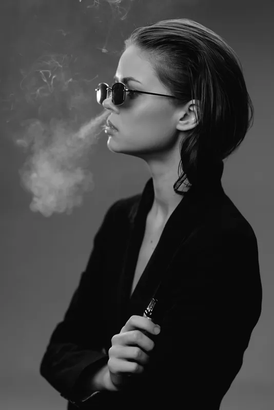 Black and white portrait of a stylish woman in sunglasses exhaling smoke, wearing dark clothing with an edgy confident look.