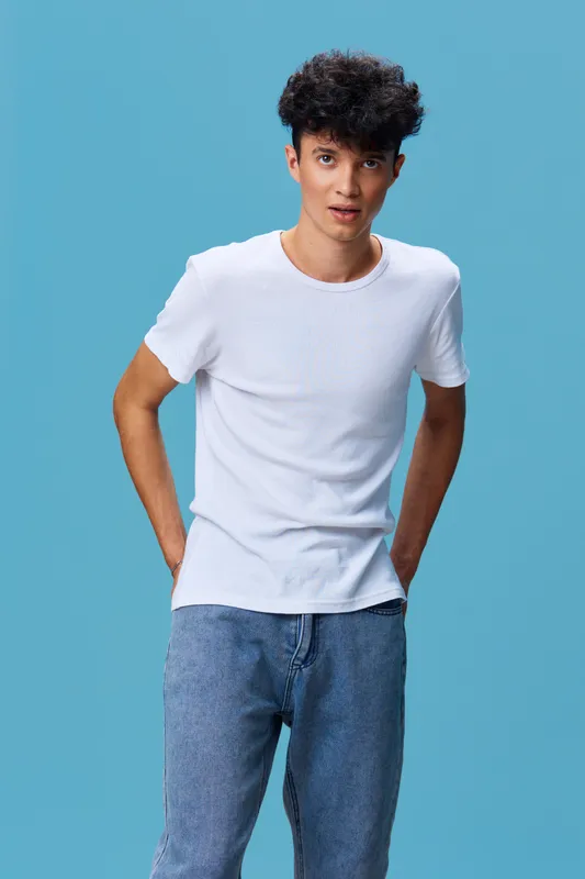 young man with curly hair wearing white t-shirt and blue jeans stands against blue background showing relaxed pose and casual style in studio lighting