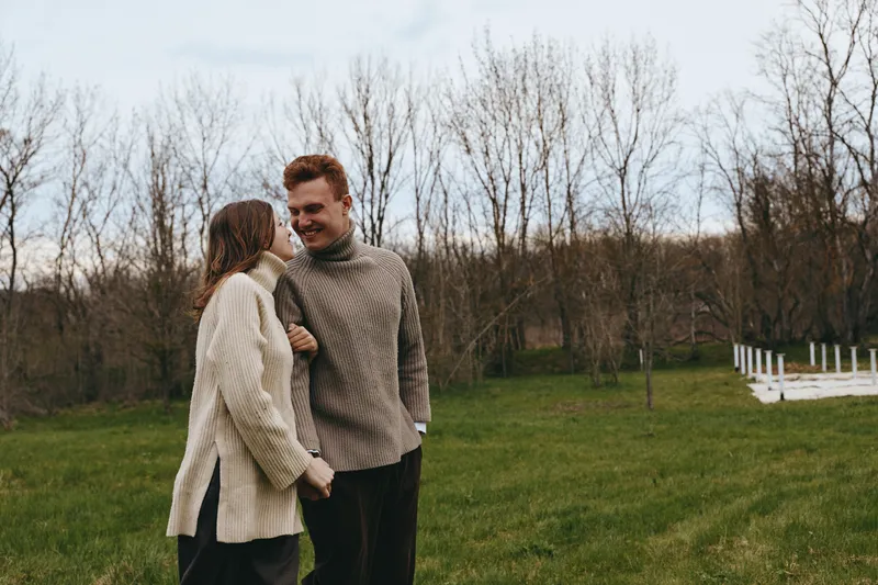 Romantic couple strolling in a grassy park on a cool day, wearing cozy sweaters, holding hands, smiling, enjoying a serene outdoor lifestyle moment amid bare trees and open field.