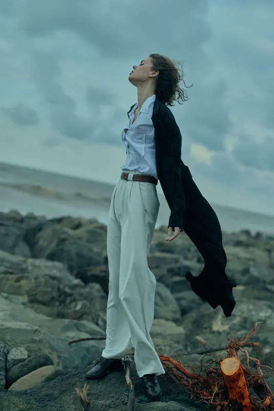 Woman in business attire stands on a windy coast, coat billowing, looking up, rugged rocks and sea behind, determined pose suggesting strength and resilience