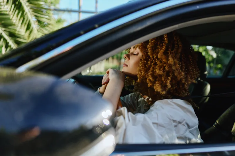 curly woman enjoying fresh air inside car, relaxed, casual style, natural light, peaceful moment, outdoor setting