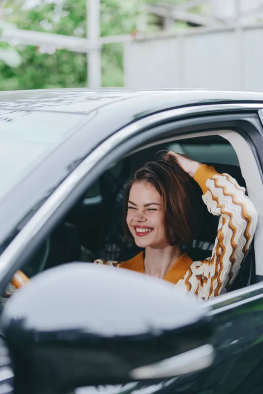 Joyful woman sits in a car with the window down, smiling brightly as sunlight fills the cabin.