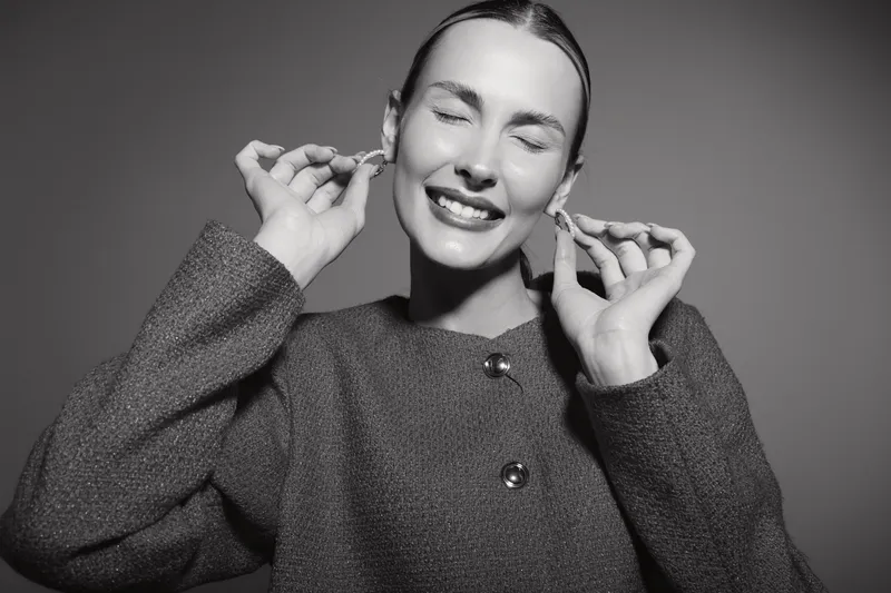 Smiling Caucasian woman with light skin and closed eyes enjoys wearing earrings in studio with soft lighting.
