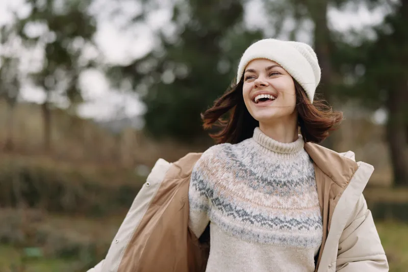 Woman enjoying an outdoor autumn breeze, smiling in a cozy sweater, beige coat and knit beanie, joyful moment in a park setting.