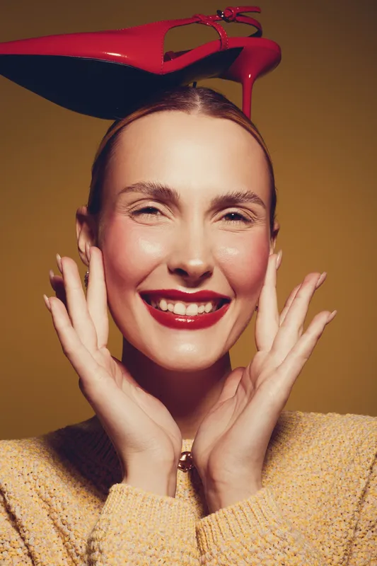 Smiling woman with red lipstick balances red high heel shoe on head, wearing yellow sweater. Studio portrait with warm lighting and playful mood, concept of fashion and joy.