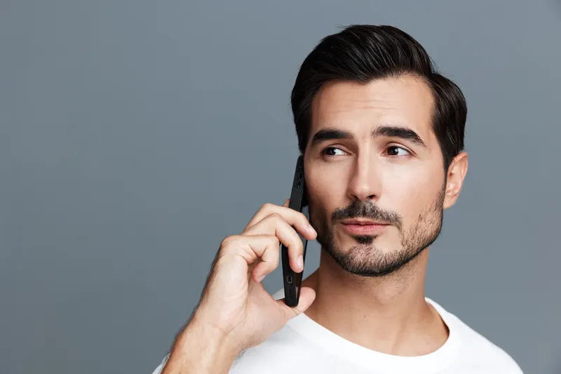 Thoughtful young caucasian man talking on mobile phone, dressed in casual white t-shirt, looking away with a confident expression against a gray background.