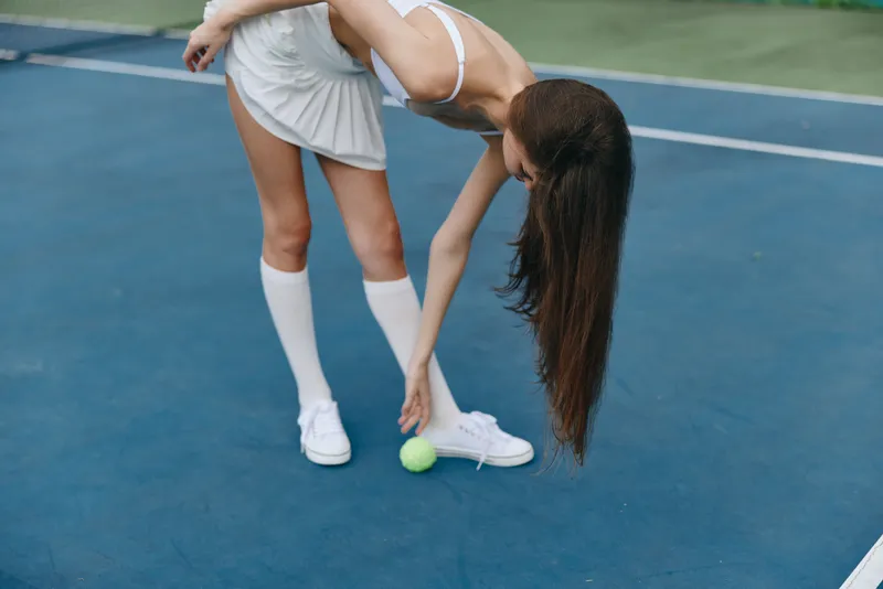 Young female tennis player in white sportswear reaching for a tennis ball on a blue court, showcasing athleticism and concentration during a match