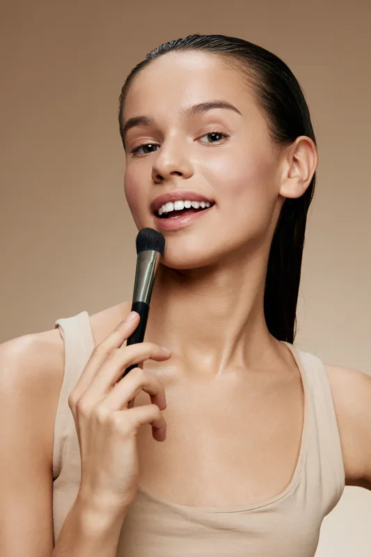Young woman smiling with a makeup brush, showcasing a natural beauty concept on a warm beige background, perfect for beauty and cosmetic themes