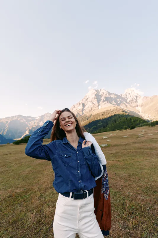 Woman with smile in denim shirt and white pants standing in mountains meadow, holding a jacket over shoulder. Outdoors portrait with natural light, alpine landscape and relaxed mood.