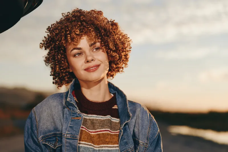 Woman with curly hair and gentle smile outdoors in golden sunset, wearing denim jacket and striped sweater, relaxed lifestyle moment, warm light, soft breeze, landscape background