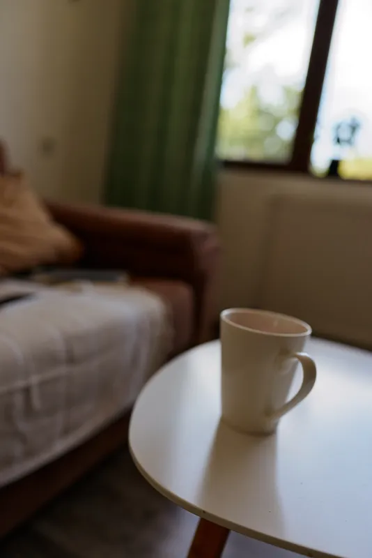 Cozy indoor scene with a white mug on a round table in a sunlit living room, blurred sofa and window in the background, calm domestic moment and simple everyday comfort