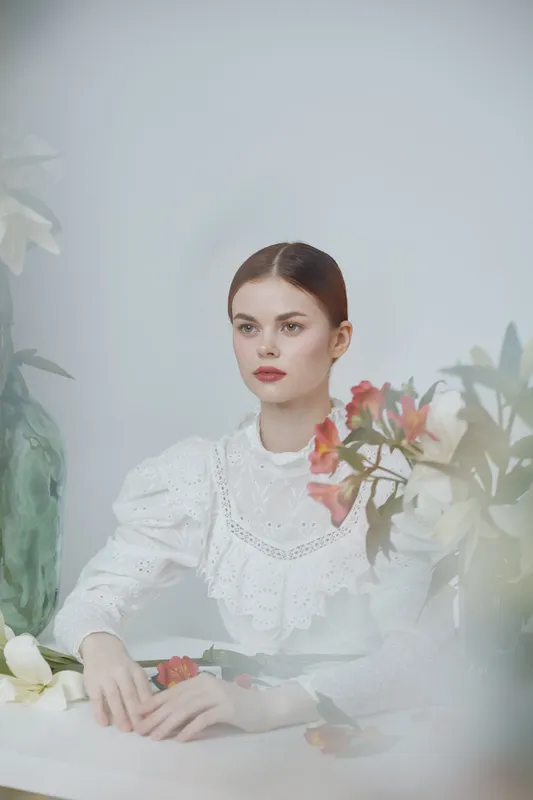 Elegant portrait of a young woman with smooth skin and red lips, wearing a vintage white dress, surrounded by soft flowers, conveying a serene and sophisticated atmosphere