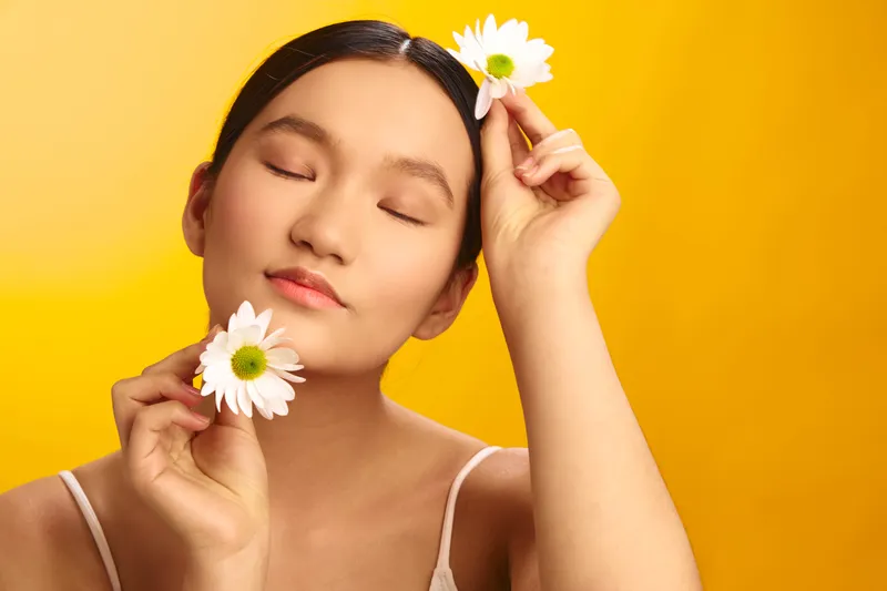 asian woman holds daisies in a serene moment against a vibrant yellow background, eyes closed, exuding calm confidence and natural beauty in a gentle, peaceful mood.