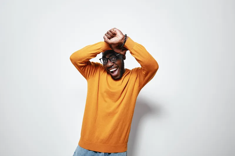 African American man with dark skin wearing glasses and orange sweater dancing happily with raised arms against white background. Casual style and joyful expression in studio.