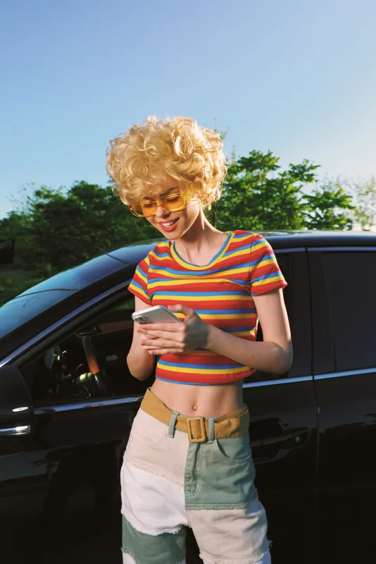Woman with curly blonde hair wearing a striped crop top and patchwork shorts stands beside a black car on a sunny day. Casual lifestyle scene with retro color, warm light, and a confident, relaxed