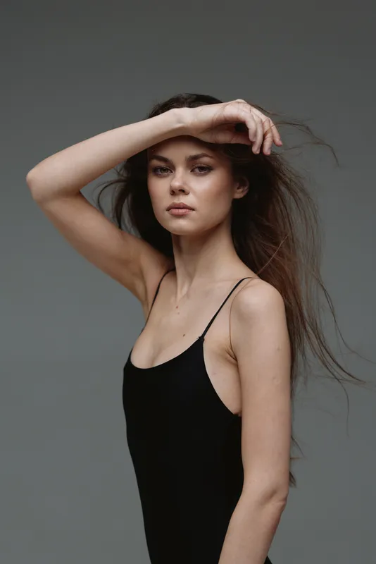Portrait of a young woman with long hair wearing a black dress posing with hand on forehead against gray background