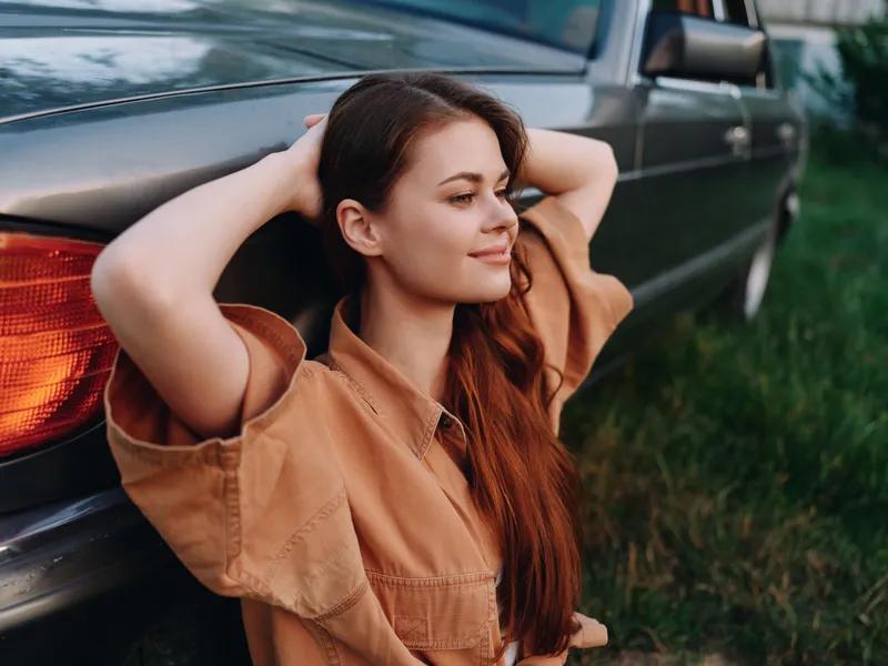 Young woman resting against a vintage car, wearing a casual brown outfit, enjoying a peaceful moment outdoors with a relaxed smile.