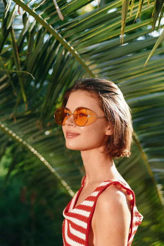 A stylish woman wearing a red and white striped top and round sunglasses stands outdoors among tropical palms. She smiles softly, enjoying sunny weather in a vibrant summer scene.