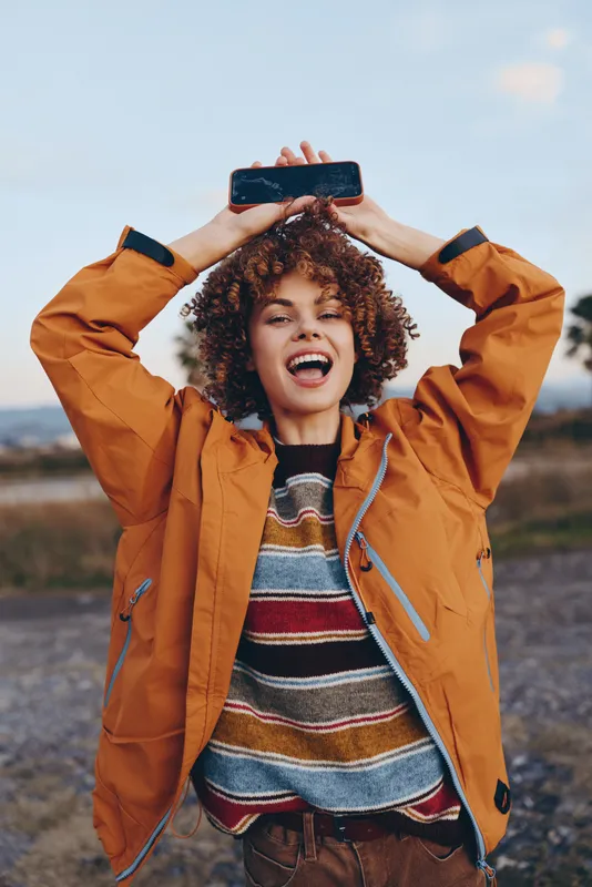 Young woman in orange jacket outdoors holds smartphone for selfie, laughing with curly hair in scenic landscape, sunny lifestyle moment, travel style snapshot.