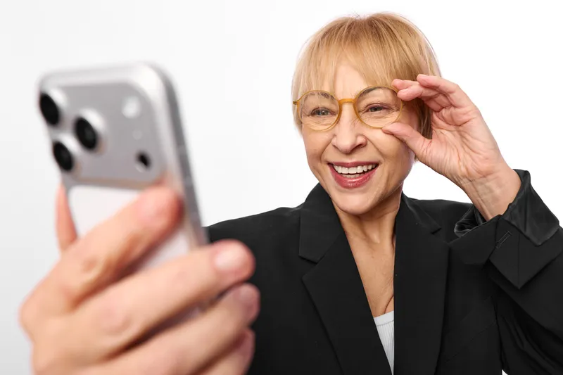 Woman selfie smartphone elderly glasses smiling portrait gesture waving hand raised, senior female with blazer, holding phone for photo, white background, closeup