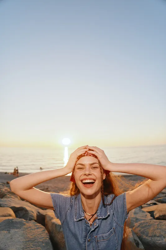 Lifestyle woman in modern boho-western denim street style laughing joyfully at sunset by the rocky shore with warm film color tones creating a vibrant mood.