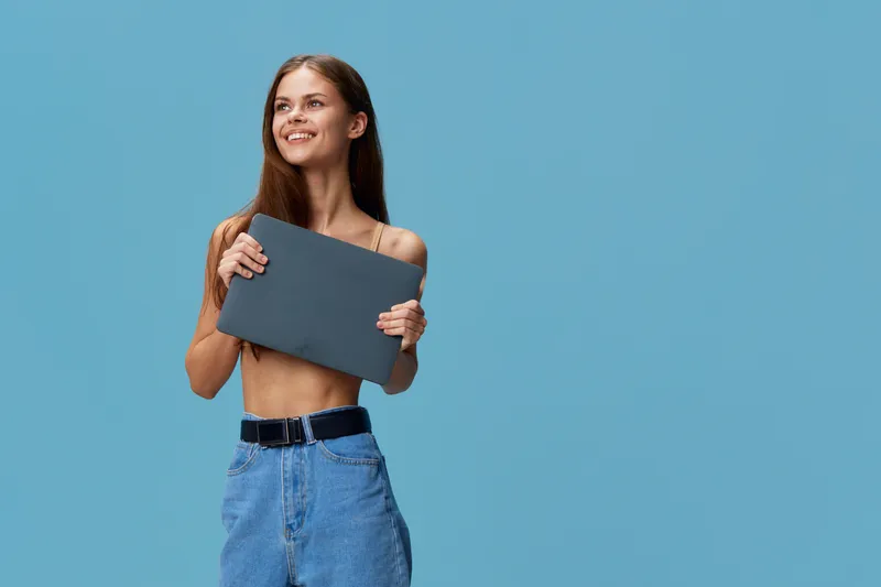 smiling woman holding laptop close to chest, casual style in blue jeans, natural light on plain blue background, youthful and positive mood, studio shot