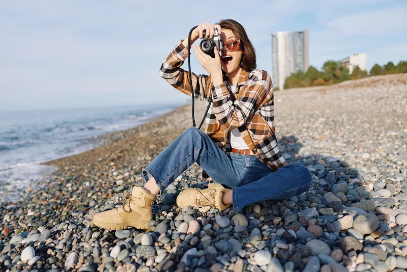 Photographer camera beach outdoor casual, sitting on a pebble shoreline, capturing coastal scenery with a handheld camera, wearing plaid shirt and jeans, boots, focused on composition and light