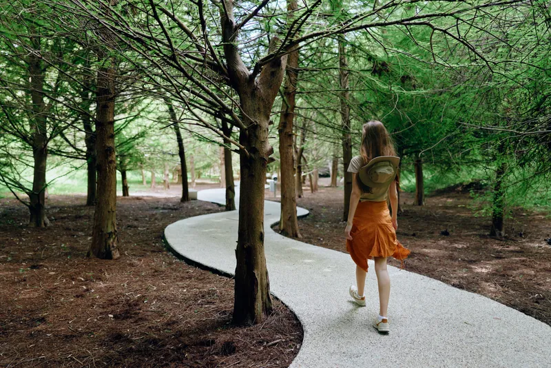 Woman walking on a winding path in a forest with backpack and trees, nature trail scene showing a young woman in orange skirt exploring shaded woodland, summer outdoor journey.