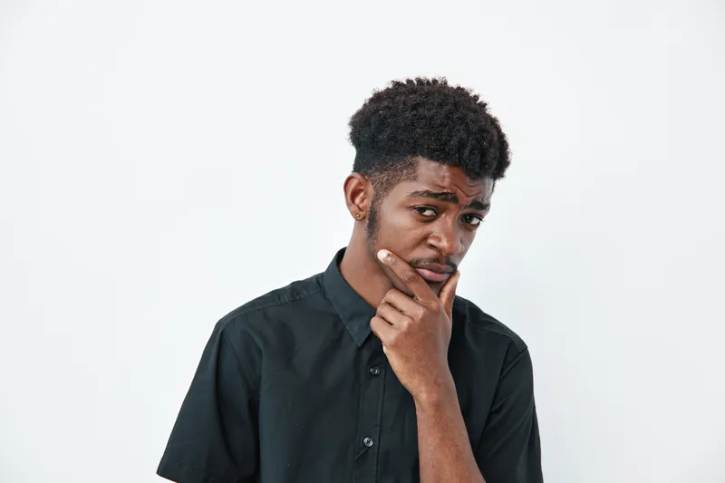 Young black man with dark skin and curly hair wearing black shirt posing thoughtful on white background. Portrait of African ethnicity male with hand on chin and serious expression.