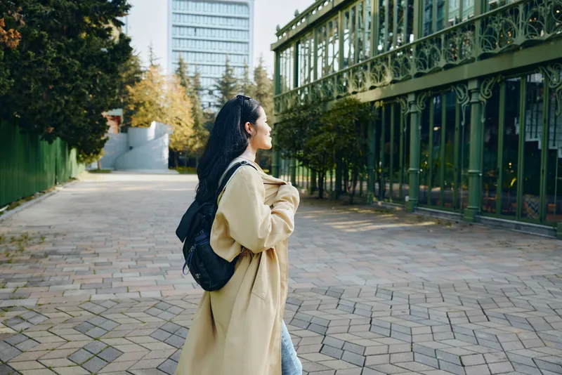 Young woman with a backpack walking along a sunny urban campus path, beige coat, glass building in the background, candid moment of exploration and daily commute beside autumn trees.