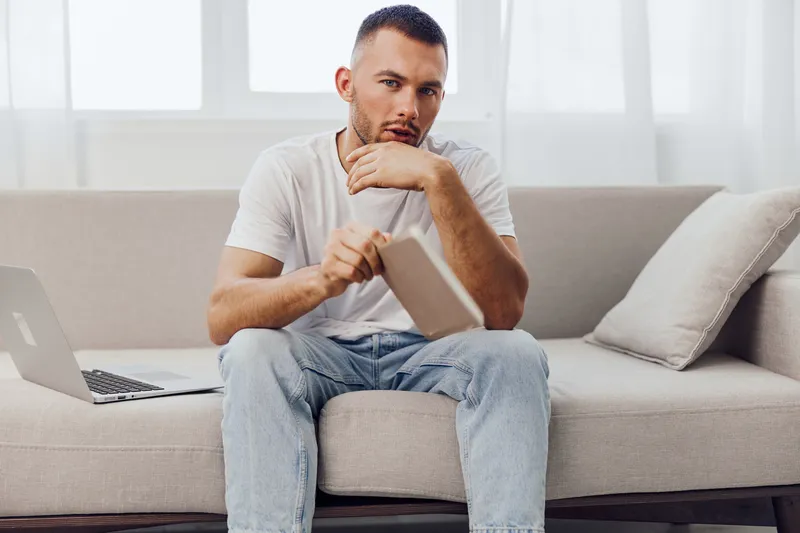 Thoughtful man seated on a sofa, holding a notebook, dressed casually in a white t-shirt, inspired by creative ideas amidst a bright and airy space.