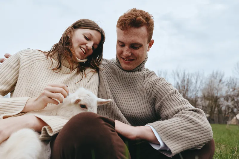 Joyful couple outdoors, smiling as they pet a small goat kid, cozy knitwear, soft breeze, sunlit grass, natural colors, serene countryside setting, relaxed interaction, candid lifestyle moment