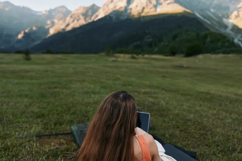 A tranquil outdoor scene shows a person lounging on a blanket and using a laptop in a wide meadow, with dramatic mountains in the distance, evoking focus and creative inspiration.