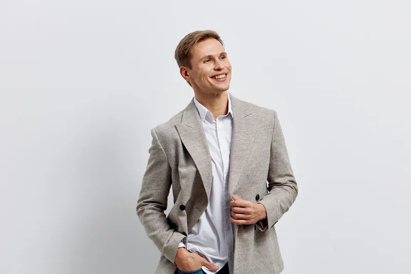 smiling young man in smart casual attire posing against a plain white background. He wears a light gray blazer and a white shirt, displaying a friendly expression and relaxed posture. Studio portrait 