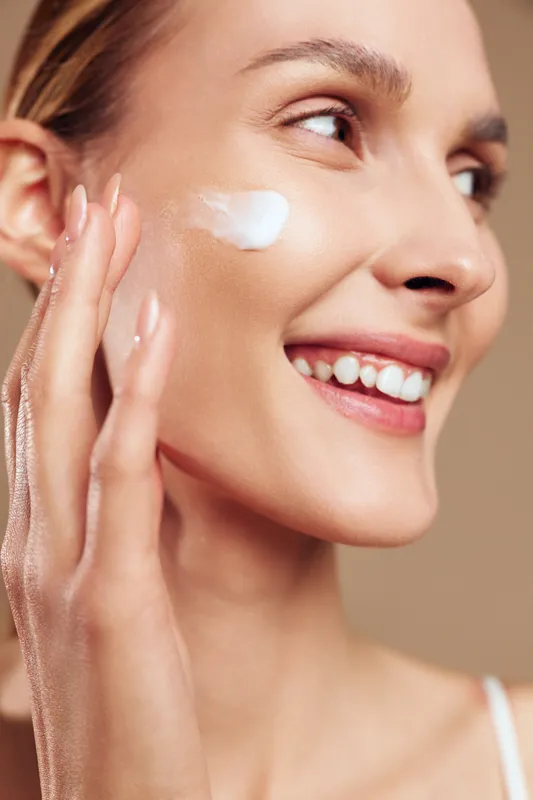 Young woman applying moisturizer cream on cheek smiling with healthy skin closeup skincare beauty routine natural glow clean fresh face
