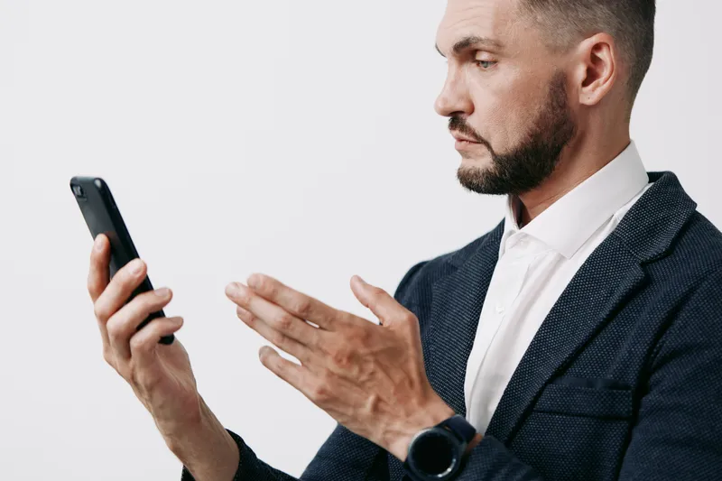 A focused business man examines a smartphone, poised and thoughtful against a solid colored backdrop, expressing concentration and subtle confidence.