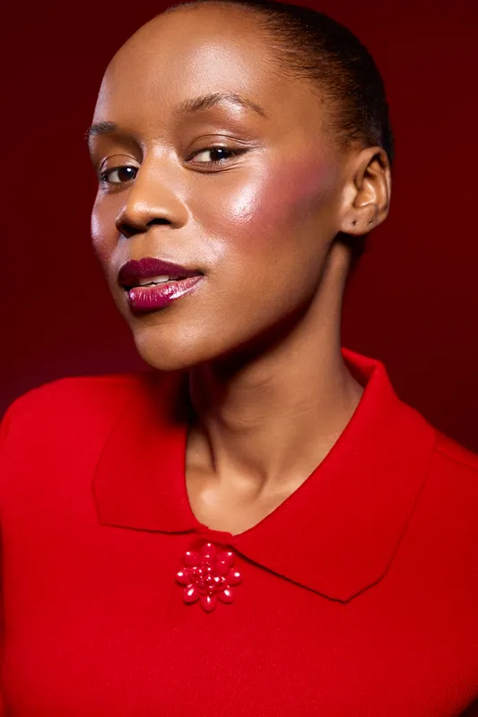 Portrait of confident black woman with dark skin wearing red dress with collar and decorative beads. Studio shot on dark red background. Concept of beauty, fashion, and elegance.