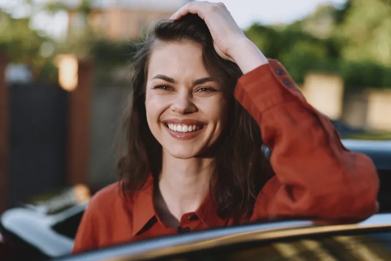 Cheerful woman leans from a car window, smiling warmly in daylight, embodying a casual lifestyle and relaxed confidence during an outdoor moment with natural light and soft breeze